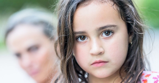 Sad Hispanic child in the foreground with mother in the background after learning about a family member's deportation.